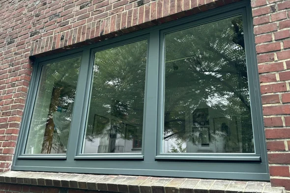 Modernes graues Fenster mit drei Scheiben in roter Backsteinmauer, reflektiert Bäume und Himmel.